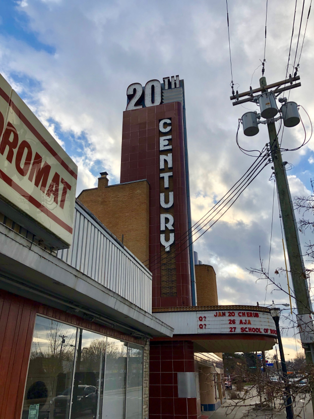 Außenansicht des 20th Century Theatre in Detroit, Michigan, mit Glaswänden und Namensschildern, umgeben von Bäumen, Strommasten und Drähten, mit einem bewölkten Himmel im Hintergrund.