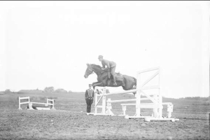 Schwarzes und weißes Foto eines Pferdes und Reiters, die über ein Hindernis springen, mit Bäumen und einem klaren Himmel im Hintergrund.