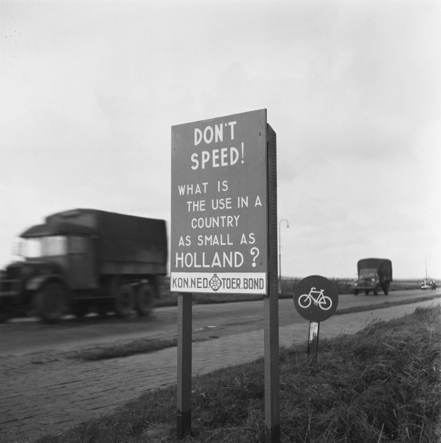 Schwarz-weißes Foto eines Lkw, der auf einer Straße mit einem "Fahren Sie nicht zu schnell"-Schild im Vordergrund und anderen Fahrzeugen, Gras, Pfählen und einem klaren Himmel im Hintergrund fährt.