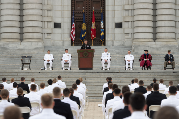 Gruppe von Menschen, die auf Stühlen vor einem Gebäude mit Säulen und einer Tür sitzen, mit einem Podium und Mikrofon in der Mitte, Fahnen im Hintergrund und einem leeren Stuhl links.