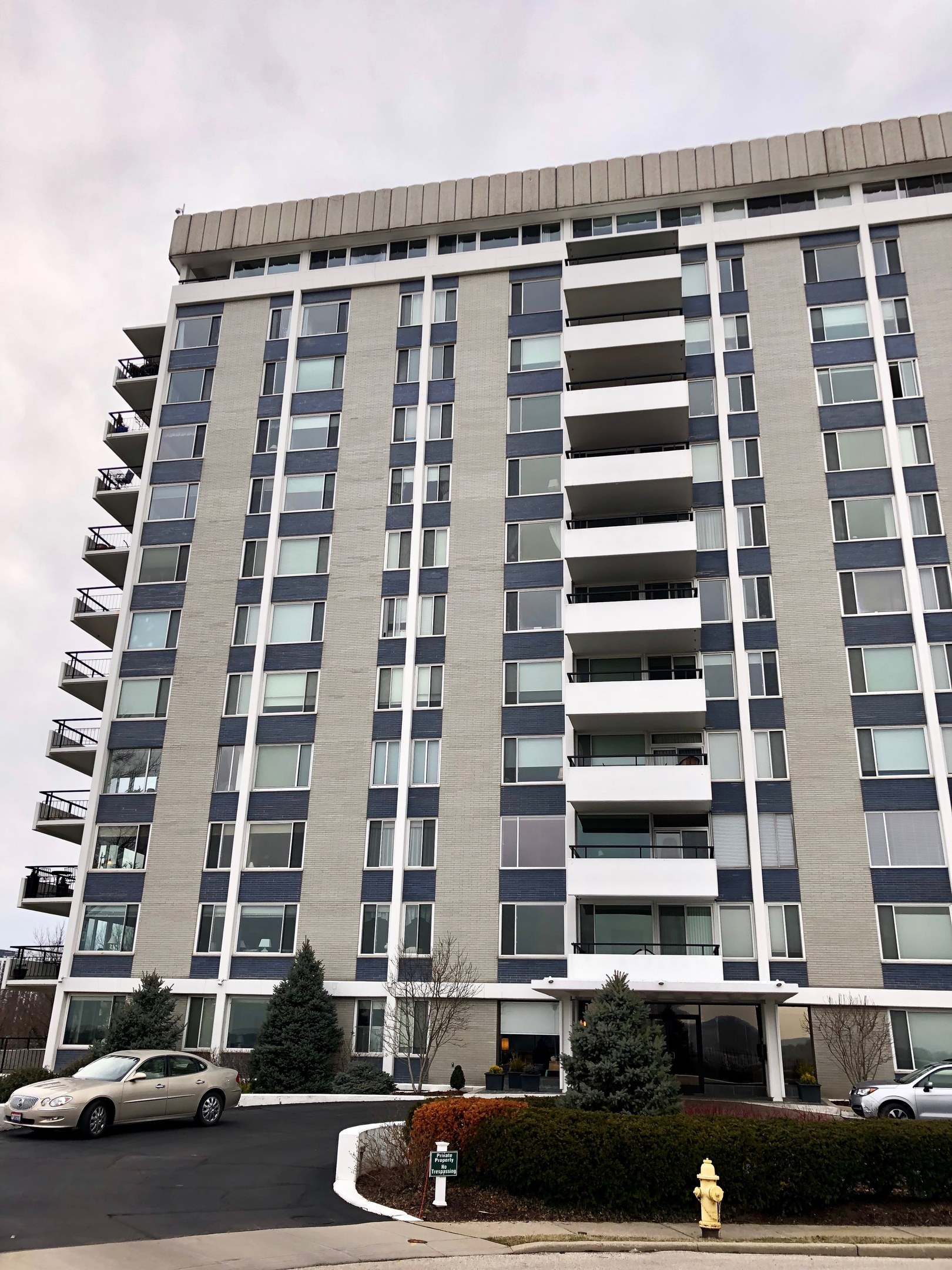 Hohes Wohnhochhaus mit zahlreichen Fenstern, einem roten Feuerhydranten im Vordergrund, Fahrzeugen auf der Straße, Grünflächen einschließlich Pflanzen und Bäumen sowie einem bewölkten Himmel im Hintergrund.