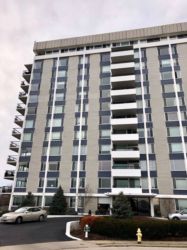 Hohes Wohnhochhaus mit zahlreichen Fenstern, einem roten Feuerhydranten im Vordergrund, Fahrzeugen auf der Straße, Grünflächen einschließlich Pflanzen und Bäumen sowie einem bewölkten Himmel im Hintergrund.