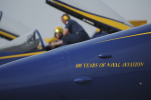 Zwei Personen sitzen im Cockpit eines blauen und gelben Kampfflugzeugs, das mit '100 Jahre der Marinefliegerei' markiert ist, vor einem klaren blauen Himmel.