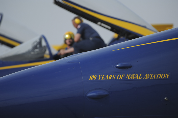 Zwei Personen sitzen im Cockpit eines blauen und gelben Kampfflugzeugs, das mit '100 Jahre der Marinefliegerei' markiert ist, vor einem klaren blauen Himmel.