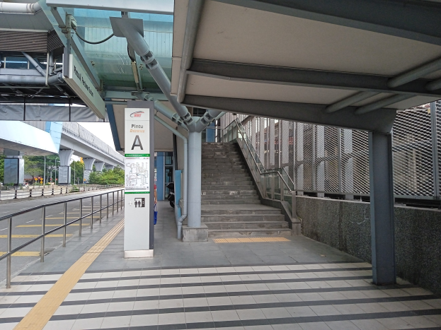 Ein Bahnsteig mit Treppe, eine Straße mit Geländer auf der linken Seite und ein Schild oben auf dem Bahnsteig, mit Bäumen, Pfählen, Gebäuden und einer Brücke im Hintergrund.