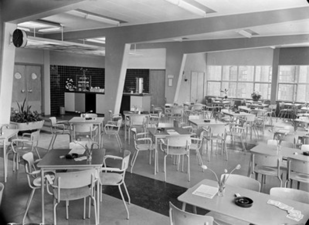 Schwarz-weißes Foto einer Cafeteria mit Tischen und Stühlen, Tellern, Besteck, Gläsern und Blumenvasen auf den Tischen sowie Türen, Fenstern und Deckenlampen im Hintergrund.