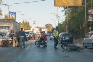 Eine Gruppe von Menschen steht um ein verunglütztes Motorrad auf der Seite einer Straße mit mehreren Fahrzeugen, darunter ein Lastwagen, und einer Hintergrundlandschaft aus Bäumen, Masten, Lichtern und Schildern unter dem Himmel.