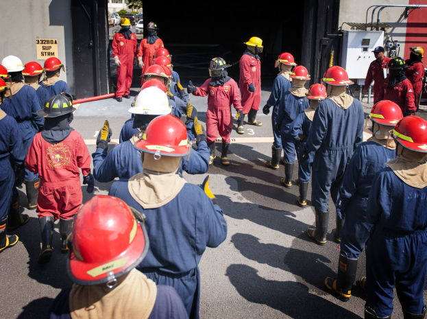 Feuerwehrleute in roter Uniform und Helmen gehen eine Straße entlang, links eine Wand mit Graffiti, Bäume und einen klaren blauen Himmel im Hintergrund.