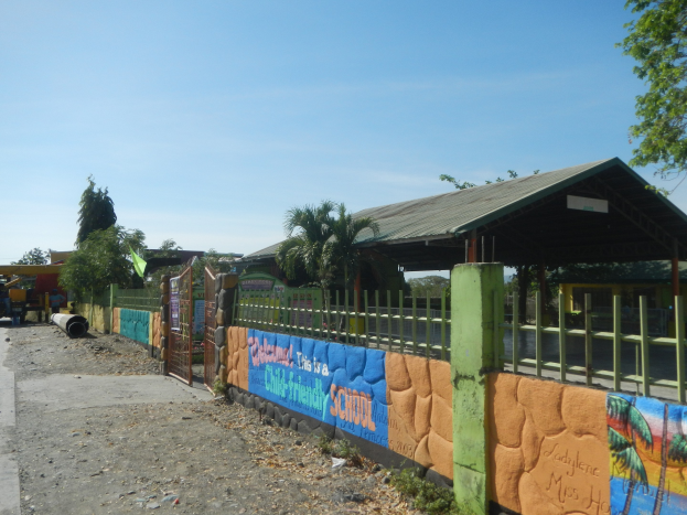 Schulgebäude mit einer Wandmalerei, umgeben von einem Zaun, einer Hütte, Bäumen, Fahrzeugen, Strommasten und einem bewölkten Himmel.