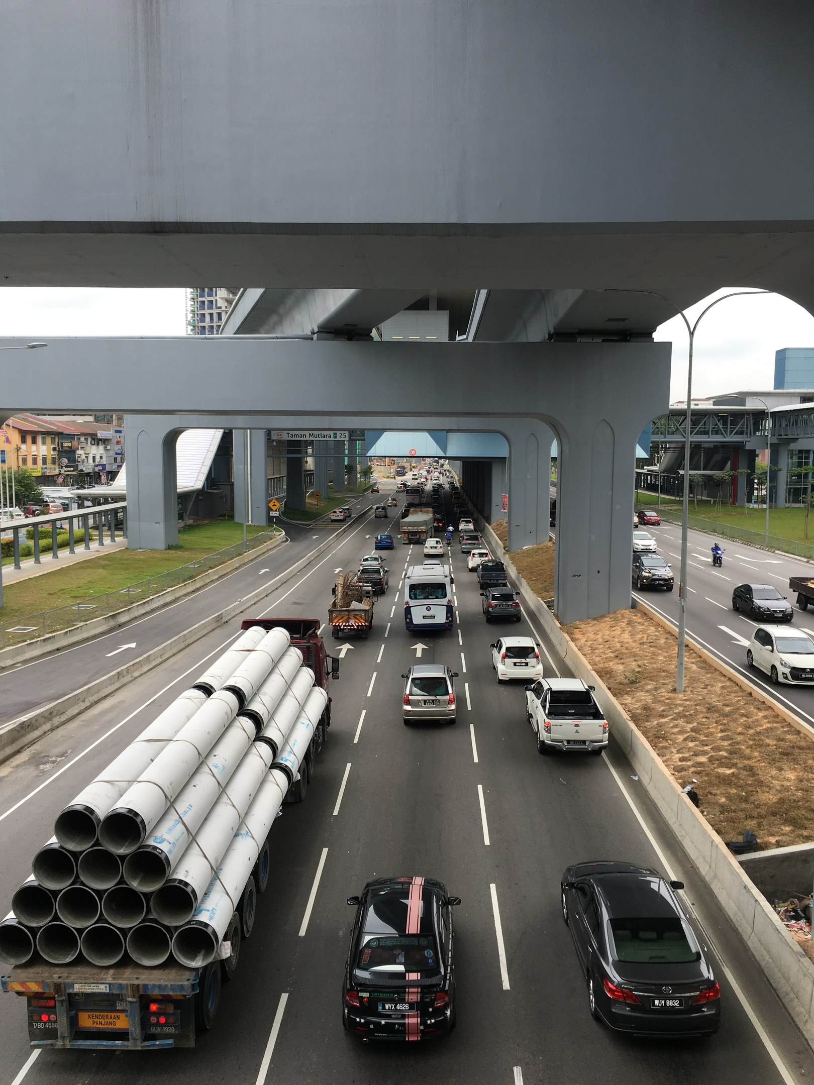Eine Autobahn mit starkem Verkehr unter einer Brücke, umgeben von Gras, Bäumen, Gebäuden, Polen und Straßenlaternen unter einem Himmel.