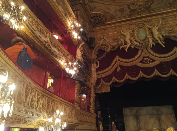 Das Innere eines Theaters mit prunkvollen Kronleuchtern, Wandskulpturen und einigen Menschen auf der Bühne, mit verstreuten Gegenständen, die eine lebhafte Atmosphäre schaffen.