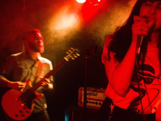 Eine Frau singt in ein Mikrofon auf der Bühne, wür ein Mann eine Akustikgitarre im Hintergrund spielt.