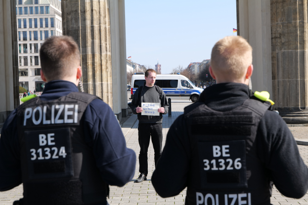 Drei Polizeibeamte stehen vor einem Gebäude, wobei ein Mann in der Mitte ein Papier hält und von Säulen und einem belebten städtischen Hintergrund flankiert wird.