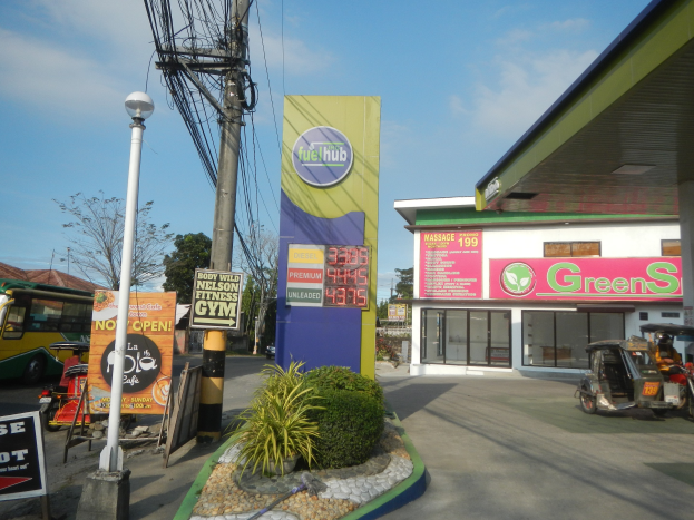 Tankstelle mit einem Schild mit der Aufschrift "Green's", Fahrzeuge auf der Straße, umliegende Pflanzen, Schilder, Pfosten, Lampen, Drähte, Gebäude, Bäume und Wolken am Himmel.