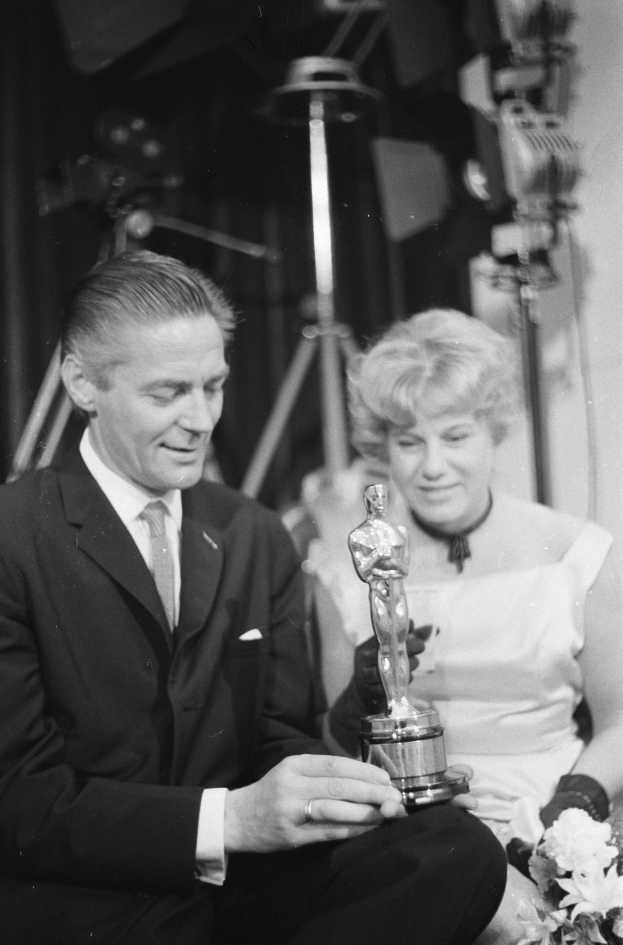 Ein Mann in einem schwarzen Blazer, weißem Hemd und Krawatte hält eine Oscar-Statue und sitzt neben einer Frau, die einen Blumenstrauß hält, beide in eleganter Kleidung, mit Lichtern, die die schwarze und weiße Szene beleuchten.