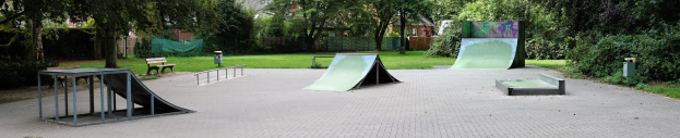 Ein Park mit einer zentralen Skateboard-Rampe, umgeben von Bänken, Geländern, Grünflächen und Gebäuden im Hintergrund.