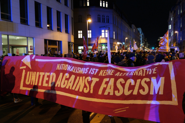 Gruppe von Menschen marschiert mit einem "Einheit gegen Faschismus"-Schild die Straße entlang, mit Gebäuden, Laternenmasten und Schildern an der Straße unter einem sichtbaren Himmel.