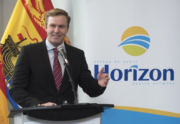 Ein Mann in einem schwarzen Blazer, weißem Hemd und roter Krawatte steht an einem Podium mit einem Mikrofon und einem Banner im Hintergrund und lächelt.