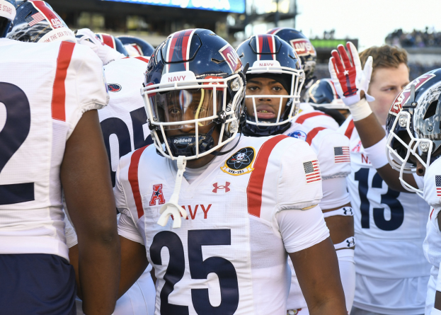 Gruppe von Männern in Football-Uniformen auf einem Feld mit Helmen und Handschuhen, Hintergrund zeigt unscharfe Zuschauer, ein Scoreboard mit "Auburn vs Virginia Tech - 11/28/15 College Football Pick, Odds, and Prediction" und Himmel.