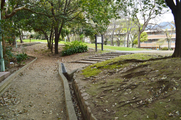 Ein Parkweg gesäumt von Bäumen und Pflanzen, flankiert von Treppen, die zu einem Gebäude führen, mit Hügeln und einem klaren blauen Himmel im Hintergrund.