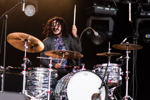 Ein Mann mit Dreadlocks, der auf der Bühne Schlagzeug spielt, hält Drumsticks mit Mikrofonständern vor sich, vor einem dunklen Hintergrund mit Bühnenbeleuchtung.