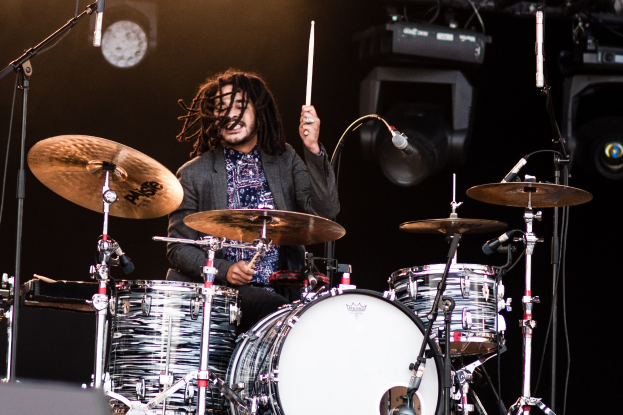 Ein Mann mit Dreadlocks, der auf der Bühne Schlagzeug spielt, hält Drumsticks mit Mikrofonständern vor sich, vor einem dunklen Hintergrund mit Bühnenbeleuchtung.