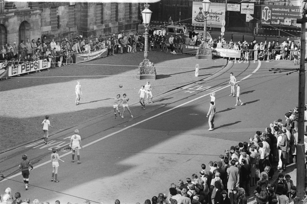 Schwarzes Bild von Menschen, die auf der Straße Fußball spielen, mit Zuschauern, Laternen, Gebäuden und Bannern im Hintergrund.