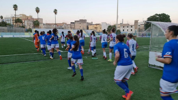 Gruppe von Frauen beim Fußballspielen auf einem Outdoor-Feld mit einem Tor und Gebäuden im Hintergrund.