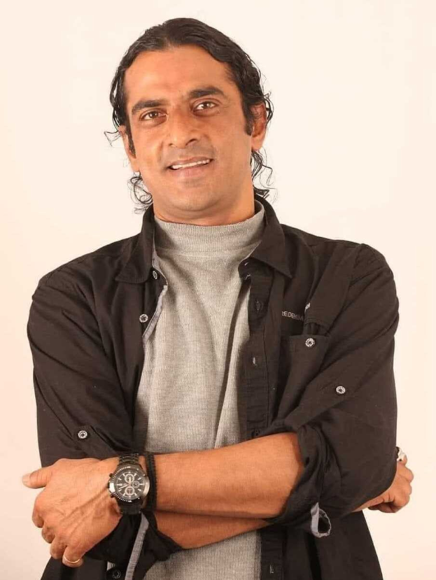 Ein Mann in einem schwarzen und aschfarbenen Kleid mit überkreuzten Armen, der eine Uhr tr├Ągt und gegen einen wei├čen Hintergrund steht, in Gedanken versunken.