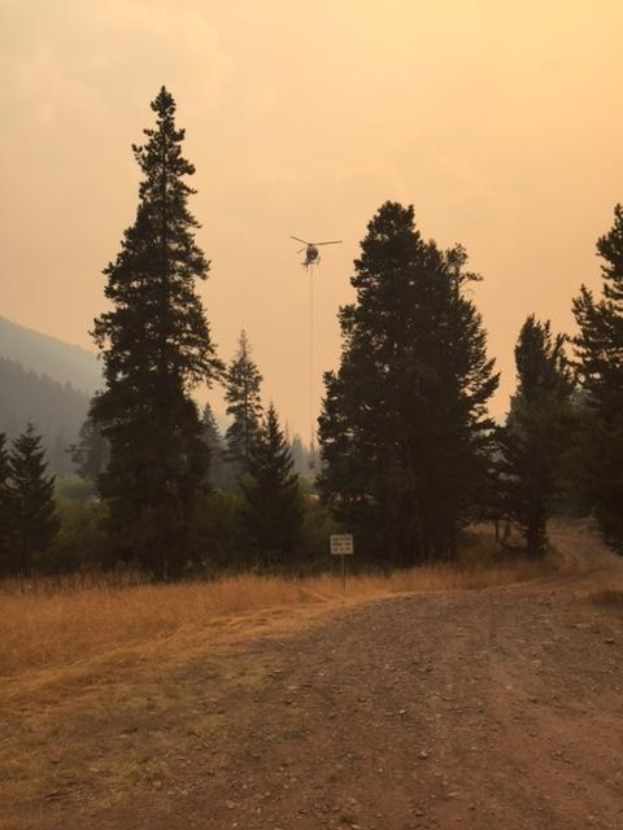 Ein Hubschrauber lässt Wasser auf einen Waldbrand fallen, während er über einen Schotterweg fliegt, mit einer Tafel an einem Pfahl im Vordergrund und Bäumen, Hügeln und einem klaren Himmel im Hintergrund.