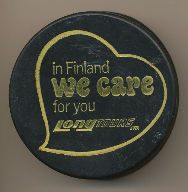 Schwarzer Hockey-Puck mit weißer Schrift "In Finland We Care For You Long Tours" auf cremefarbenem Hintergrund.