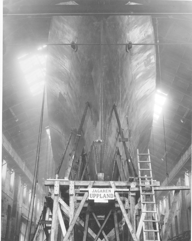 Ein Schwarz-Weiß-Foto eines großen Boots im Bau in einer Werft, das eine Leiter, Holzpfosten, ein Schild, Metallrahmen, Deckenlampen und Fenster zeigt.