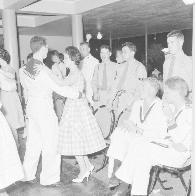 Schwarzes und weißes Bild von Militärpersonal, das auf einer Tanzfläche tanzt, einige sitzen und einige stehen, mit Säulen und Lichtern im Hintergrund.