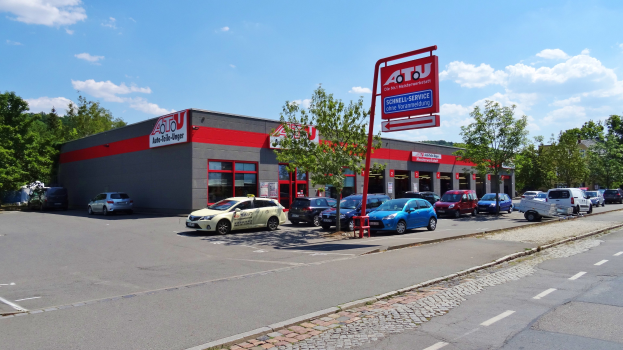 Ein Autohaus mit mehreren geparkten Autos vorne, umgeben von Gebäuden, Bäumen, Strommasten, Schildern und einer Straße unter einem bewölkten Himmel.