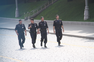 Drei Polizisten in Uniform und Mützen, die eine Straße entlanggehen mit Pfählen, Geländern, Treppen und Gras im Hintergrund.