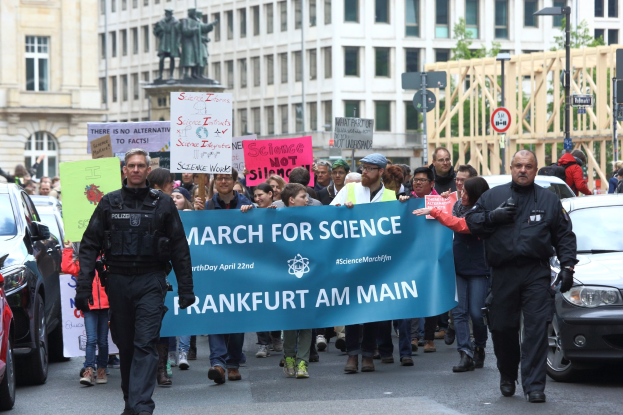 Gruppe von Menschen marschiert mit einem "March for Science Frankfurt am Main"-Schild die Straße entlang, während Autos daneben fahren und Gebäude, Statuen, Laternenpfähle, Schilder und Bäume im Hintergrund zu sehen sind.