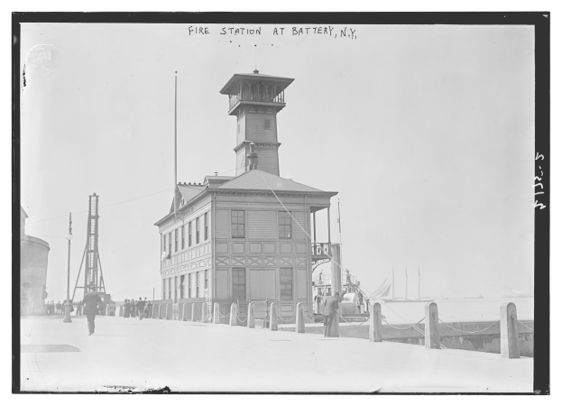 Schwarze und weiße Aufnahme eines Feuerwagens in Battery, New York, mit einem Gebäude, Menschen, Pfählen, einem Zaun, Booten auf dem Wasser und einem bewölkten Himmel.