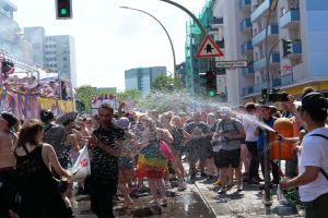 Menschen auf einer Christopher Street Day Parade, die sich gegenseitig mit Wasser bespritzen und Gegenstände halten, mit einem Banner links und Gebäuden, Bäumen und Straßenelementen im Hintergrund.