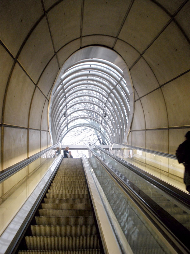 Rolltreppe in einer U-Bahn-Station mit Menschen darauf, flankiert von Wänden und oben von einem Dach abgeschlossen.