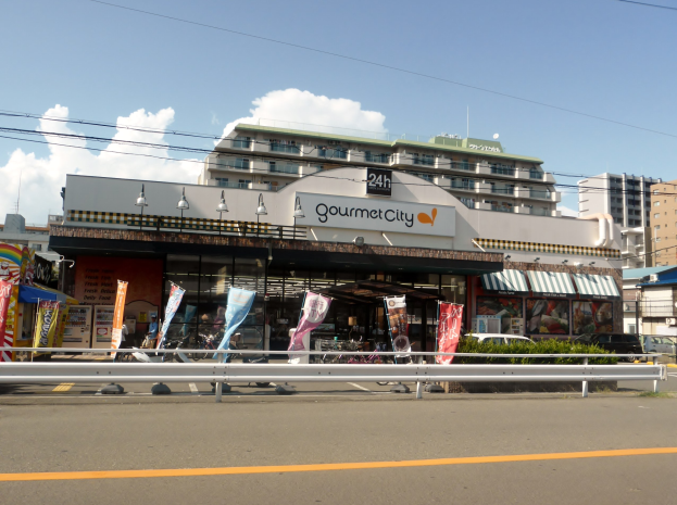 Eine belebte Stadtstraße mit hohen Gebäuden, Straßenlaternen, Werbetafeln, Fahrzeugen, Absperrpollern, Grünflächen und einem bewölkten Himmel, mit einem Restaurant in der Mitte, das verschiedene Speisen anbietet.