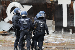 Gruppe von Polizisten in schwarzen Uniformen und blauen Helmen, die vor einem mit Graffiti bedeckten Gebäude während einer Straßenschlacht stehen, mit verstreuten Papieren und Gegenständen auf dem Boden.