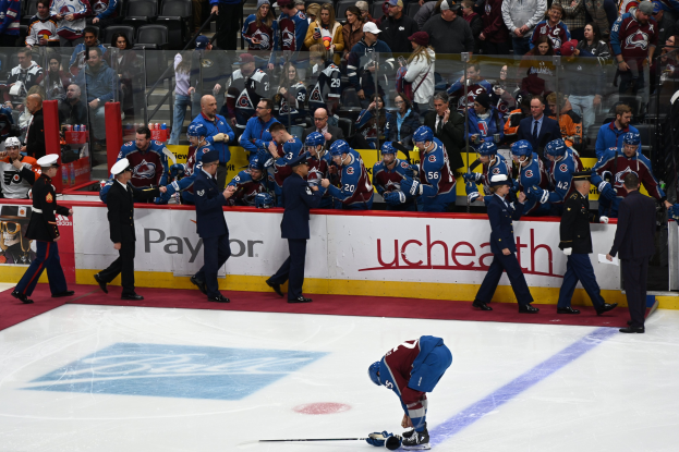 Hockey player on ice surrounded by teammates and opponents, with spectators in background during Colorado Avalanche vs San Jose Sharks game on 3/24/15.