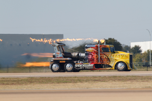 Ein Lkw mit Flammen, die aus der Rückseite austreten, f├Ąhrt auf einer Stra├če mit Gras, einem Zaun, B├Ąumen, einem Geb├Ąude, einem Stra├čenpfahl, Dr├Ąhten und einem bew├Âlktem Himmel, wobei ein Reifen und ein Rad zu sehen sind.