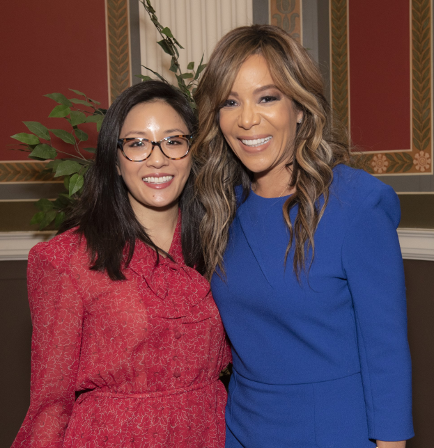 Zwei Frauen stehen nebeneinander, lächeln, vor einer Wand mit einer Pflanze und einem Vorhang, bei der Oscar-Verleihung 2019 für Frauen im Film; die Frau rechts trägt ein blaues Kleid, die Frau links trägt ein rotes Kleid und eine Brille.