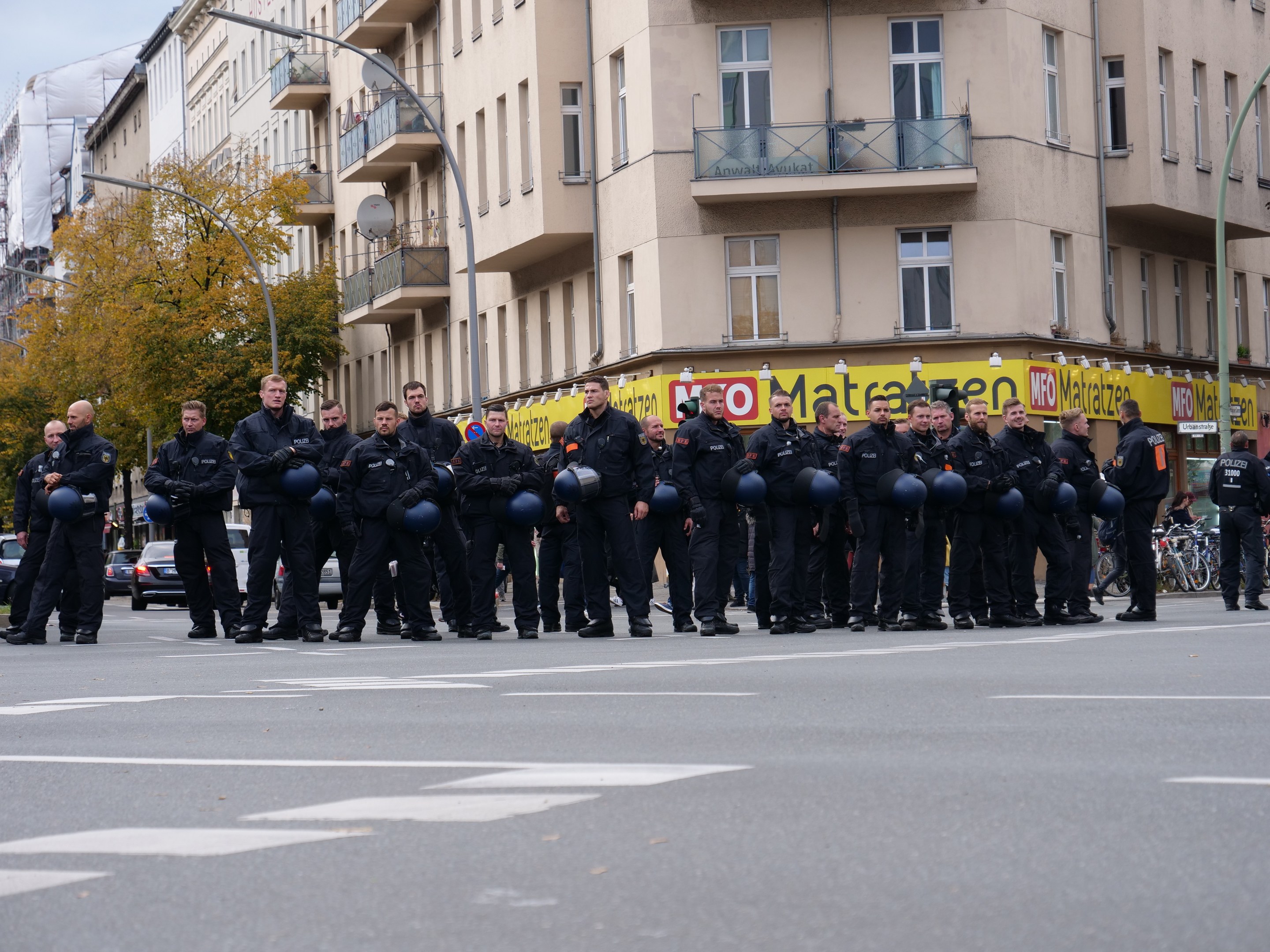 Eine Gruppe von Polizisten in schwarzen Uniformen mit blauen Helmen steht in einer Straße, die von Laternenmasten, Bäumen und Gebäuden mit Glasfenstern und Balkonen gesäumt ist, unter einem klaren blauen Himmel.