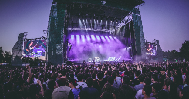Eine große Menge an einem Musikfestival schaut auf einer beleuchteten Bühne mit Instrumenten und Bildschirmen, die vor einem Gebäude, Bäumen und einem blauen Himmel aufgebaut sind.