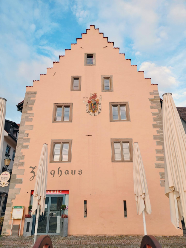 Ein rosafarbenes Gebäude mit einem zentralen Uhrenturm, umgeben von Fenstern, Schirmen, einem Schild, einer Straßenlaterne, Topfpflanzen und einem bewölkten Himmel in München, Deutschland.