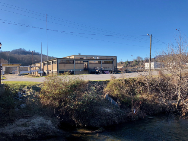 Grünes Feld mit einem Fluss, der durch es fließt, gesäumt von Bäumen, Pflanzen, Gras, Steinen und Straßeninfrastruktur, mit einem Gebäude im Hintergrund unter einem klaren blauen Himmel, an einem geplanten Wasseraufbereitungsanlagenstandort.