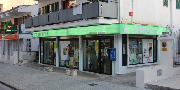 Eine Straßenecke mit einer Apotheke und benachbarten Gebäuden, sichtbarer Beschilderung, Strommasten, Fahrzeugen und Topfpflanzen unter einem klaren blauen Himmel.