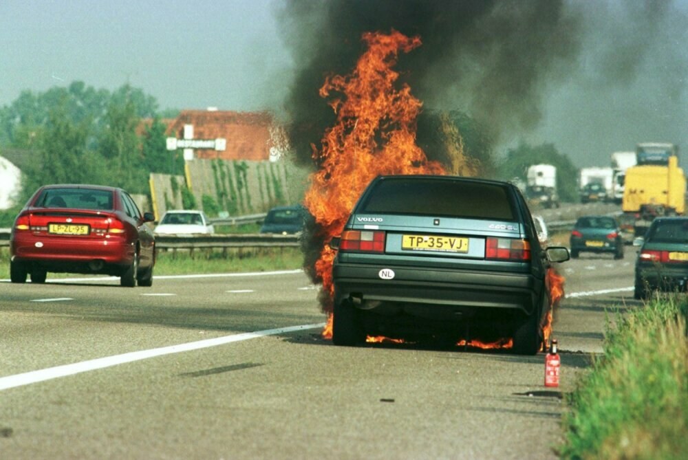 Ein Auto, das in Flammen steht, am Straßenrand mit anderen Fahrzeugen in der Nähe, umgeben von Bäumen, Gebäuden und einem klaren blauen Himmel, mit Gras und einem Feuerlöscher auf der rechten Seite.
